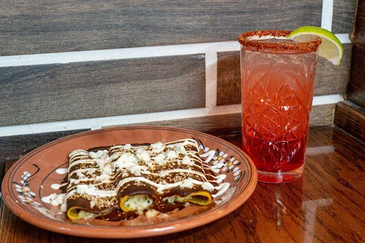 Enchiladas drizzled with mole and crema on a hand-painted clay plate, next to a tall red cocktail with a chili rim and a lime wedge