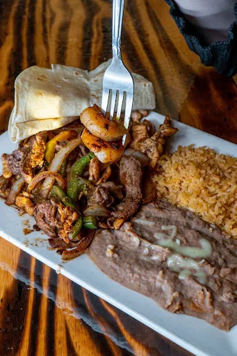 Fajitas plate with grilled steak, chicken and shrimp over peppers and onions, served with Mexican rice, refried beans and flour tortillas on a wooden table