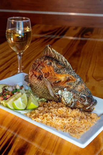 Mojarra frita: a whole fried tilapia served with Mexican rice, lime wedges and pico de gallo on a wooden table, beside a glass of white wine